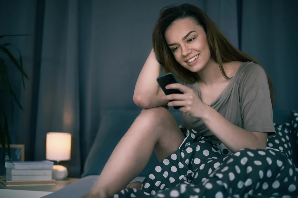 Hôtesse souriante sur son lit, la nuit, téléphone en main